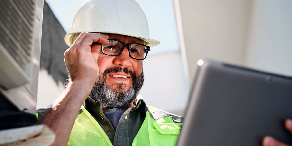 SAFEbuilt inspector stands on construction site in white hat and yellow vest reviewing inspection notes on iPad