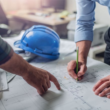 Hands near hard hat point at paper plan review while another man writes with pencil